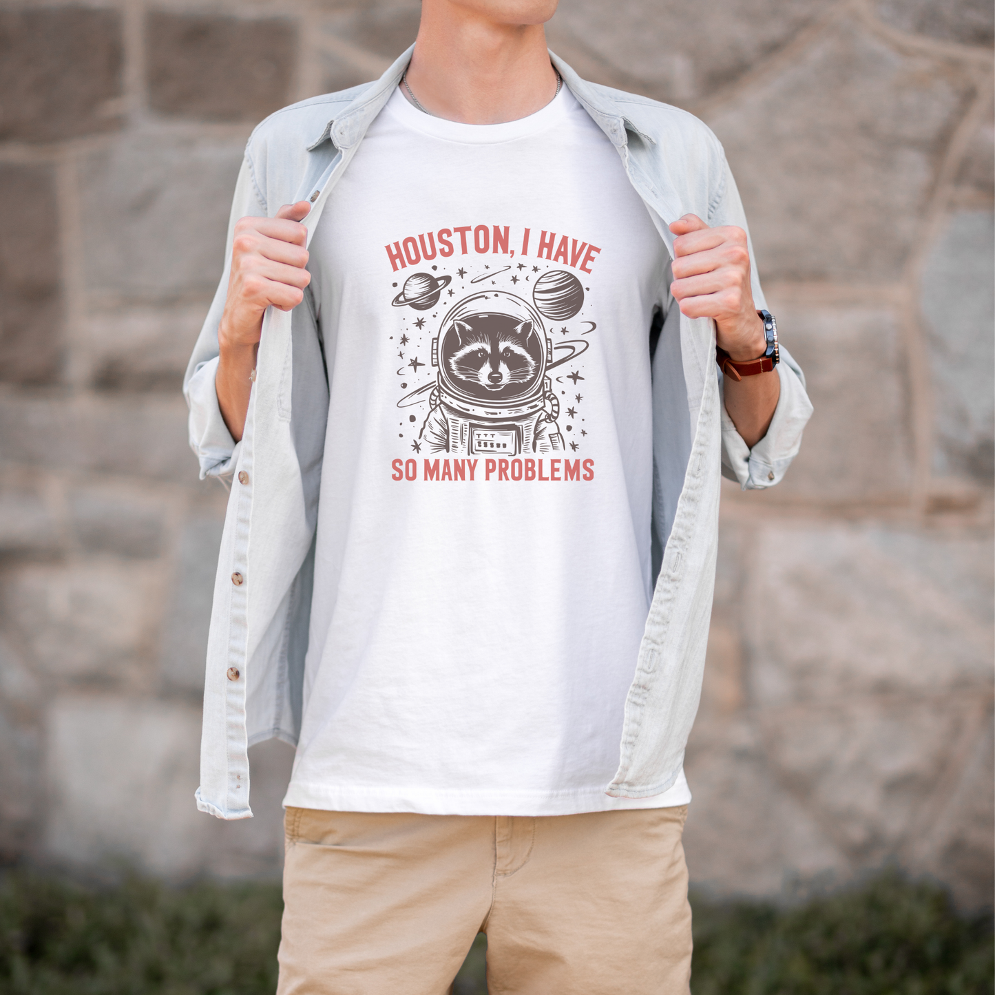 Person wearing a white t-shirt with a graphic design and text, standing against a stone wall.houston i have so many problems, raccoon astroanut