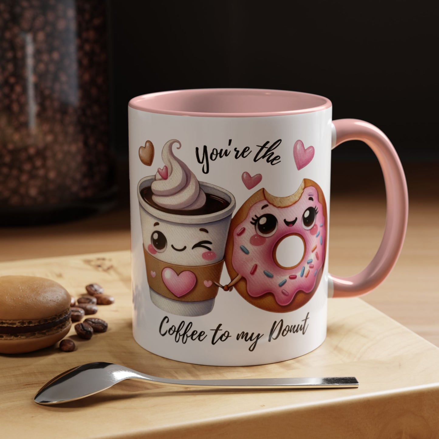 Mug with cartoon coffee cup and donut, text 'You're the coffee to my donut', on a wooden surface with coffee beans and a spoon.