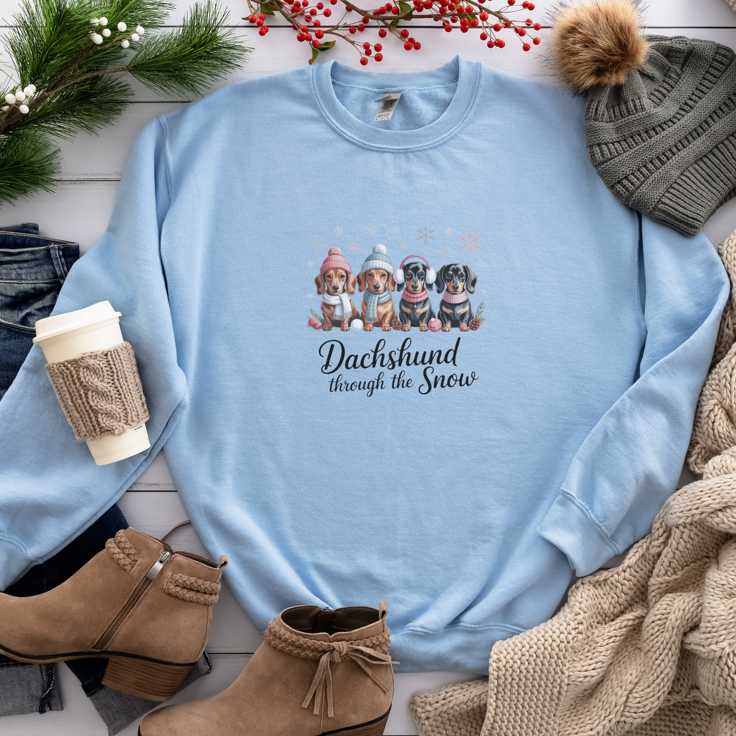 Light blue sweatshirt with dachshund design and text, surrounded by winter accessories on a wooden surface. dachshund through the snow