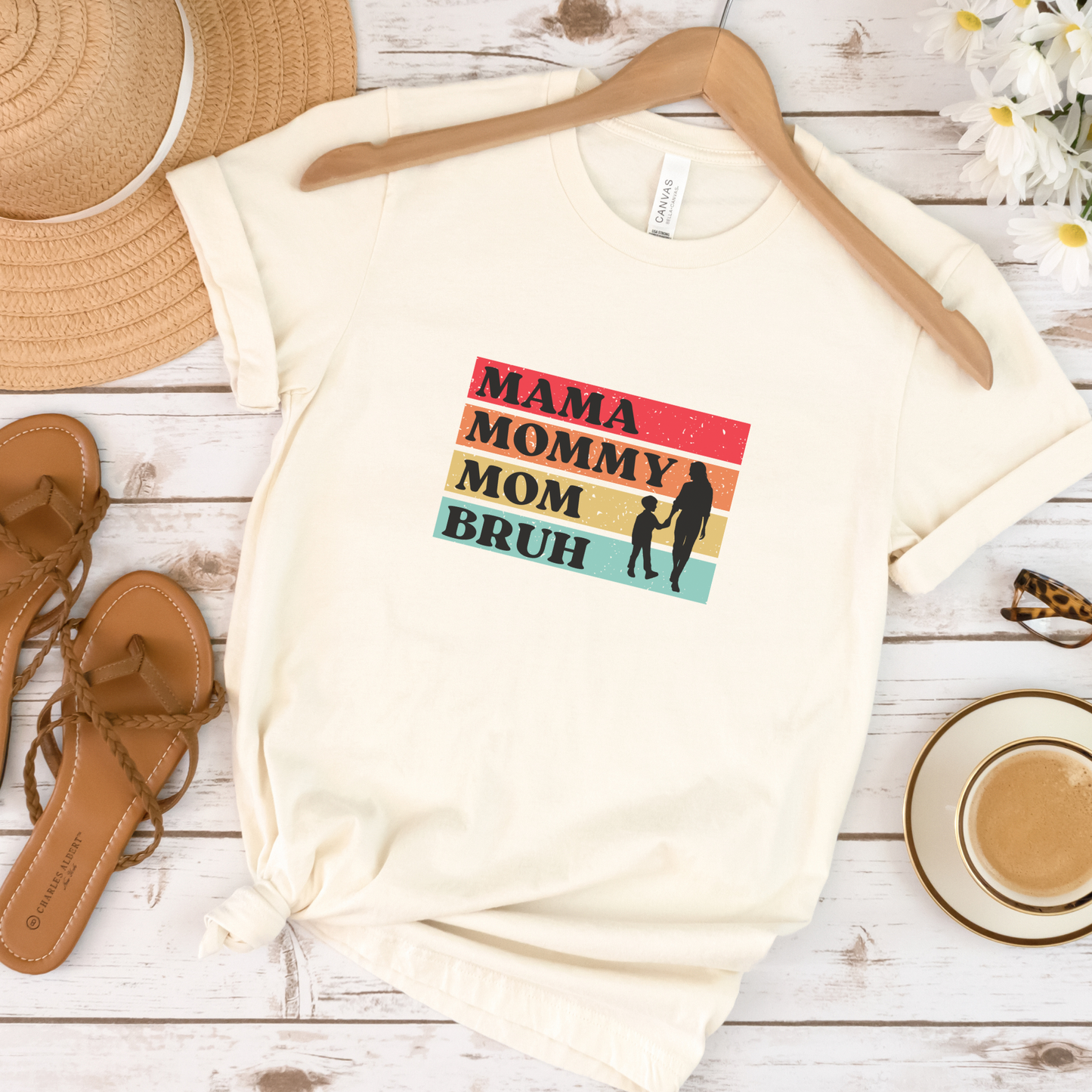 Beige t-shirt with 'Mama Mommy Mom Bruh' graphic on a wooden surface with accessories.