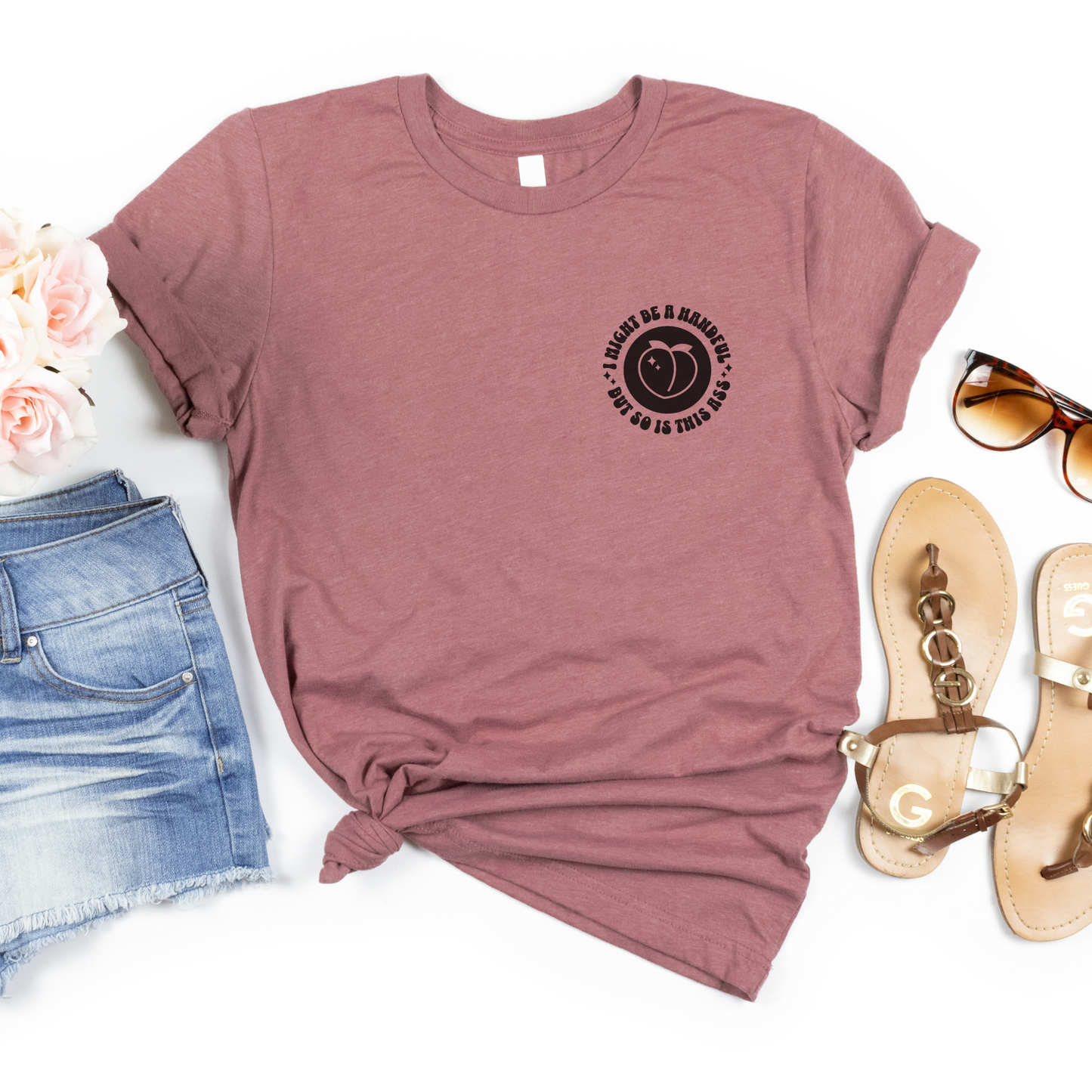 Mauve t-shirt with a graphic design, sunglasses, sandals, and denim shorts on a white background,  I might be a handful but so is this ass