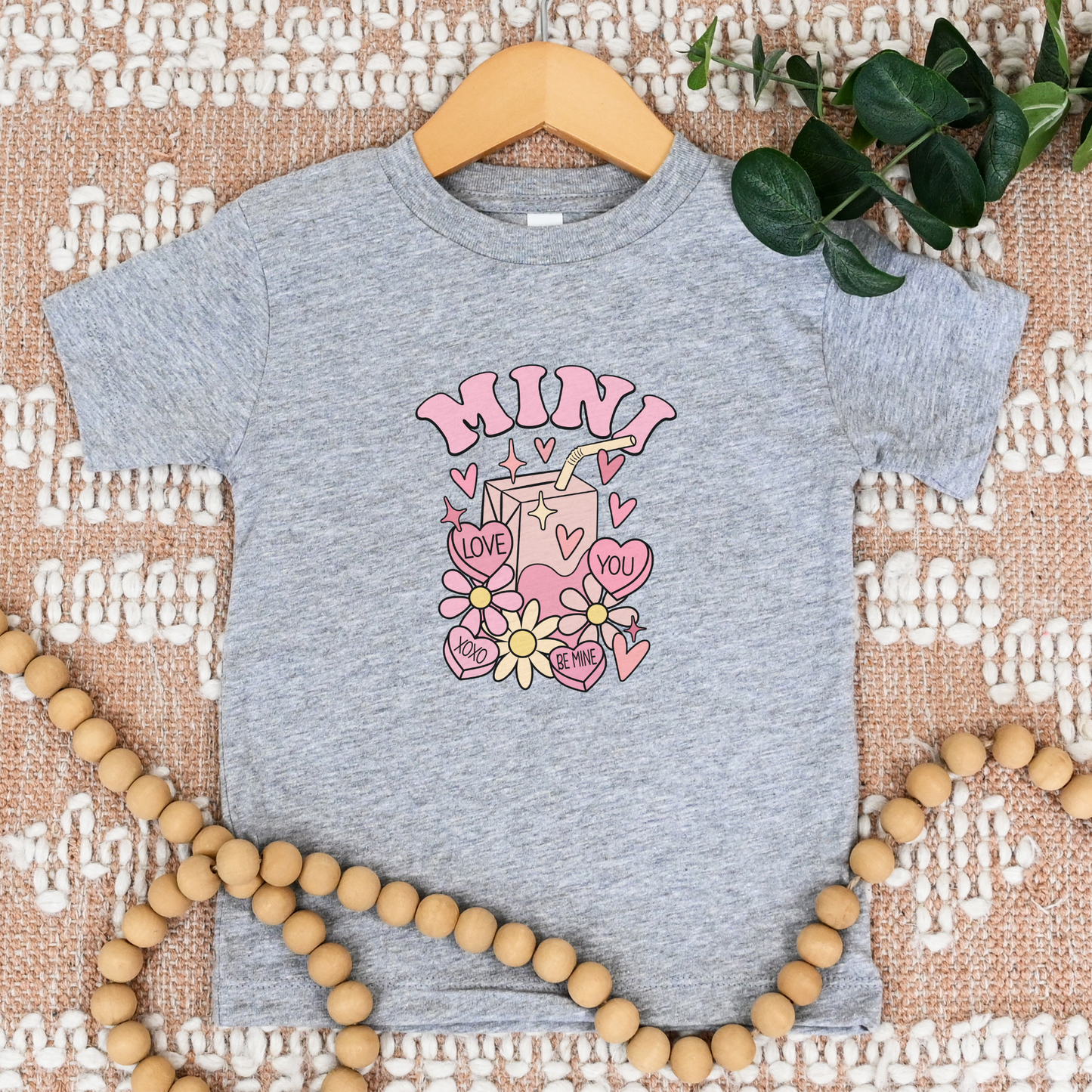 Gray t-shirt with pink graphic design on a textured surface with wooden beads and green leaves. valentines mini 