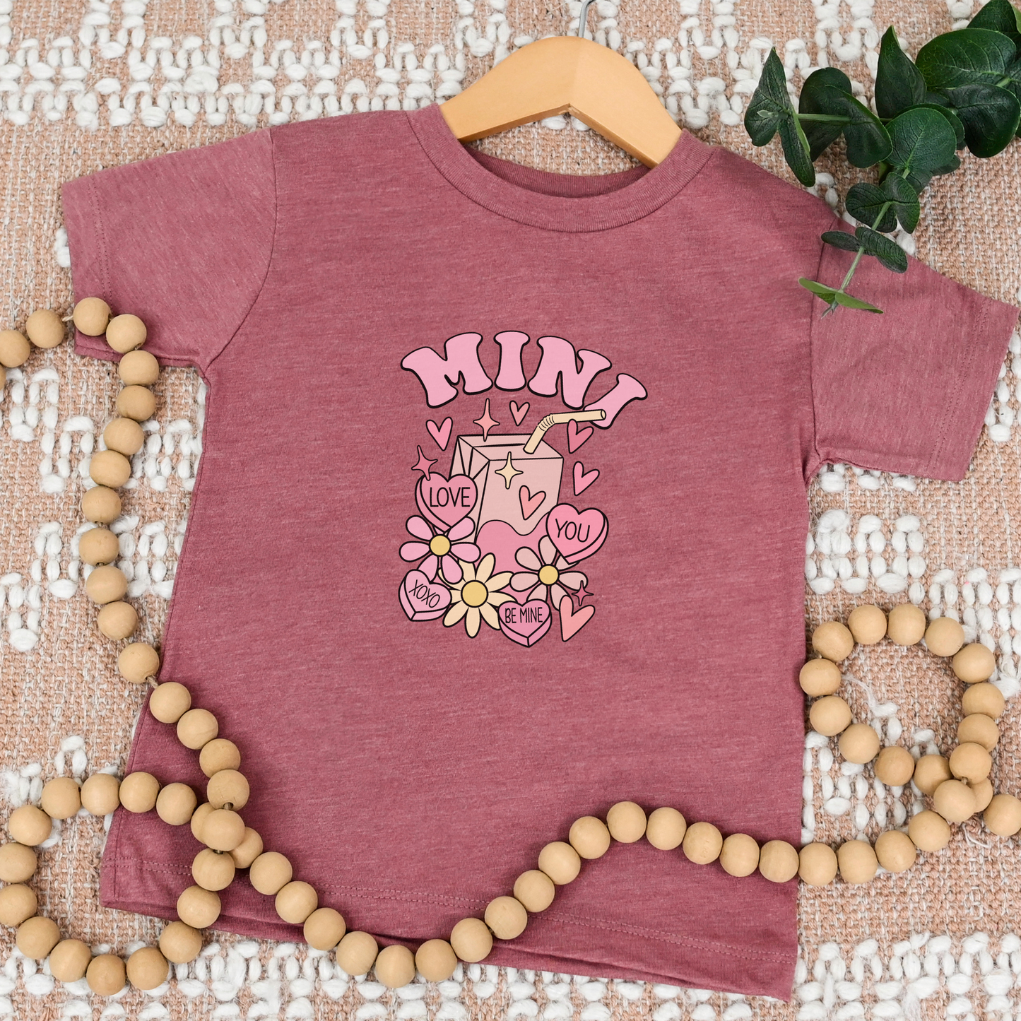 heather mauve tee with 'MINI ' design on a textured surface with wooden beads and green leaves. mama mini matching tee