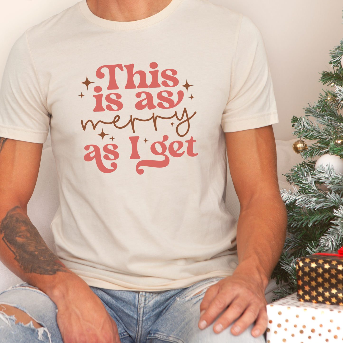 Person wearing a white t-shirt with 'This is as merry as I get' text, sitting next to a decorated Christmas tree.