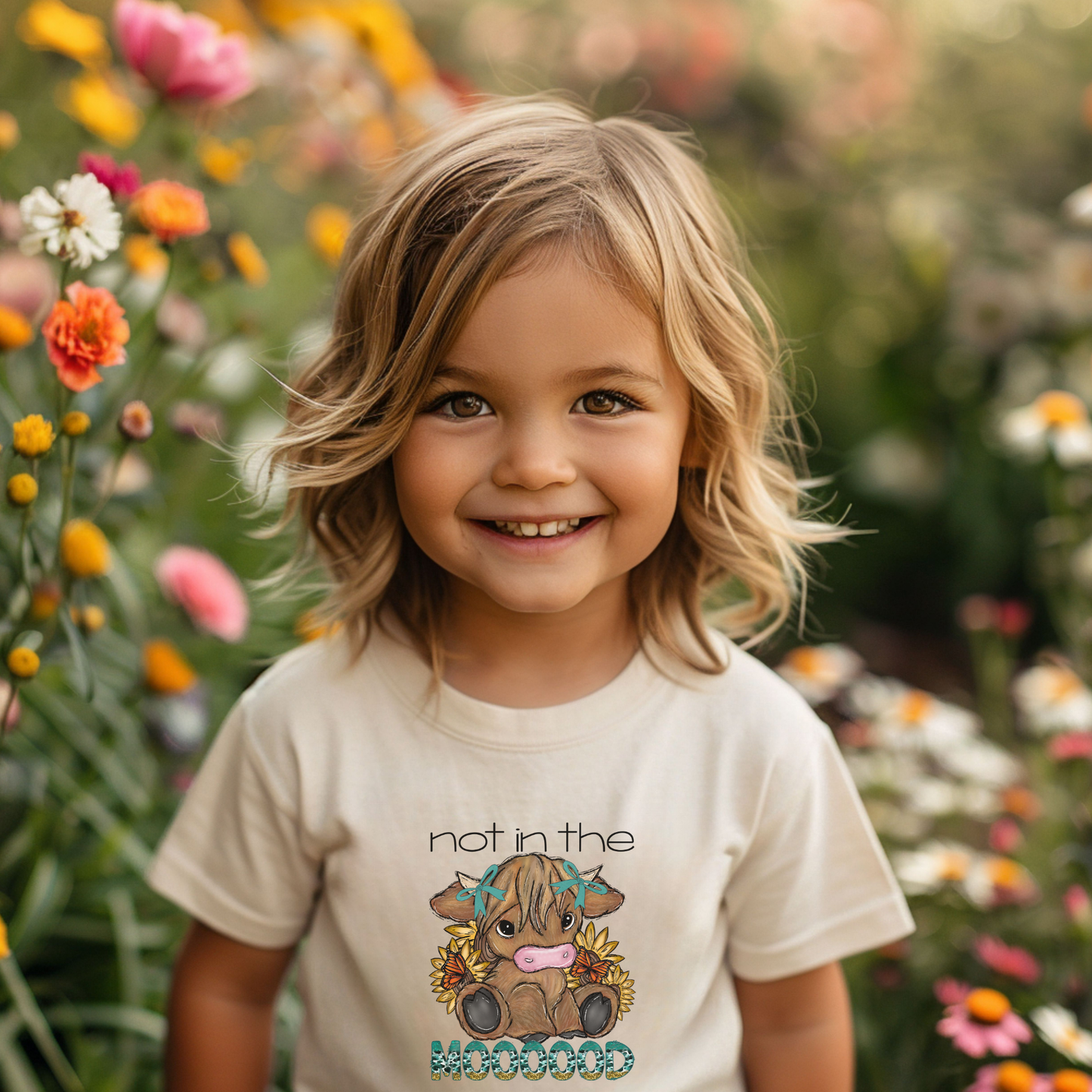 Child wearing a t-shirt with a graphic and text, standing in a garden with flowers. not in the mood baby highland cow