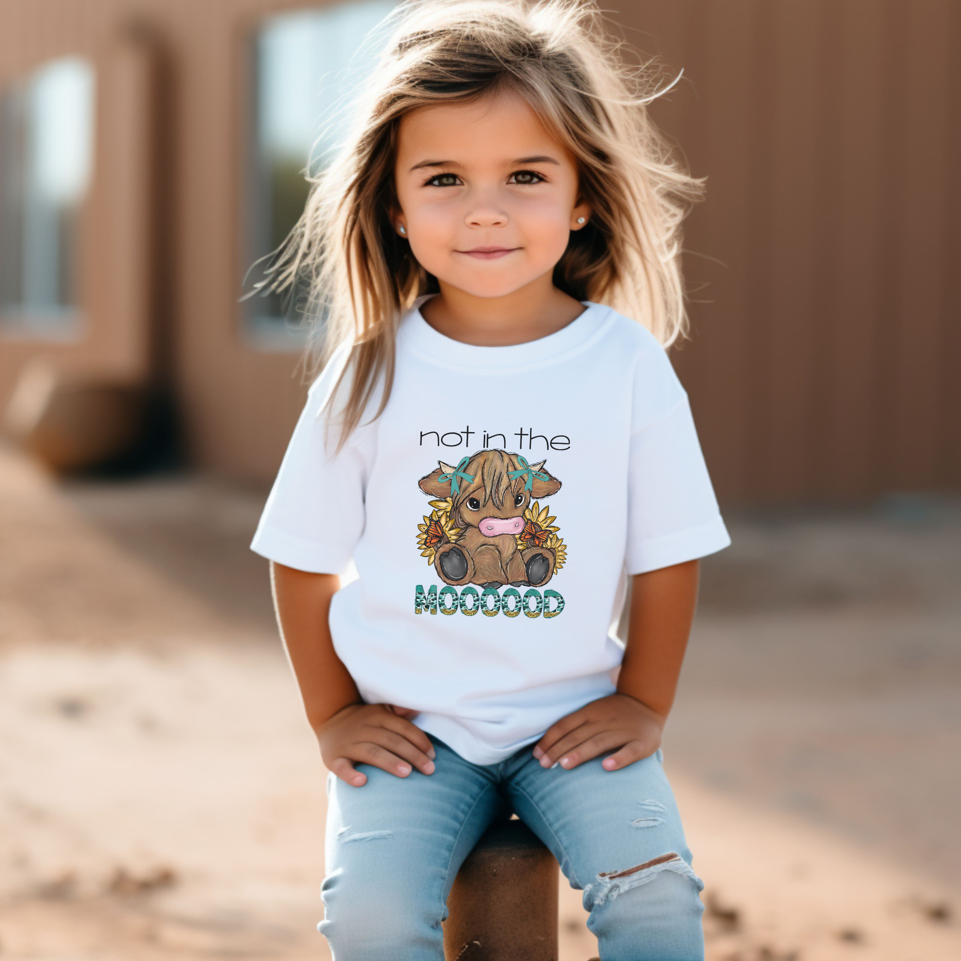 Child wearing a white t-shirt with a graphic design and text, sitting outdoors. not in the mood baby highland cow