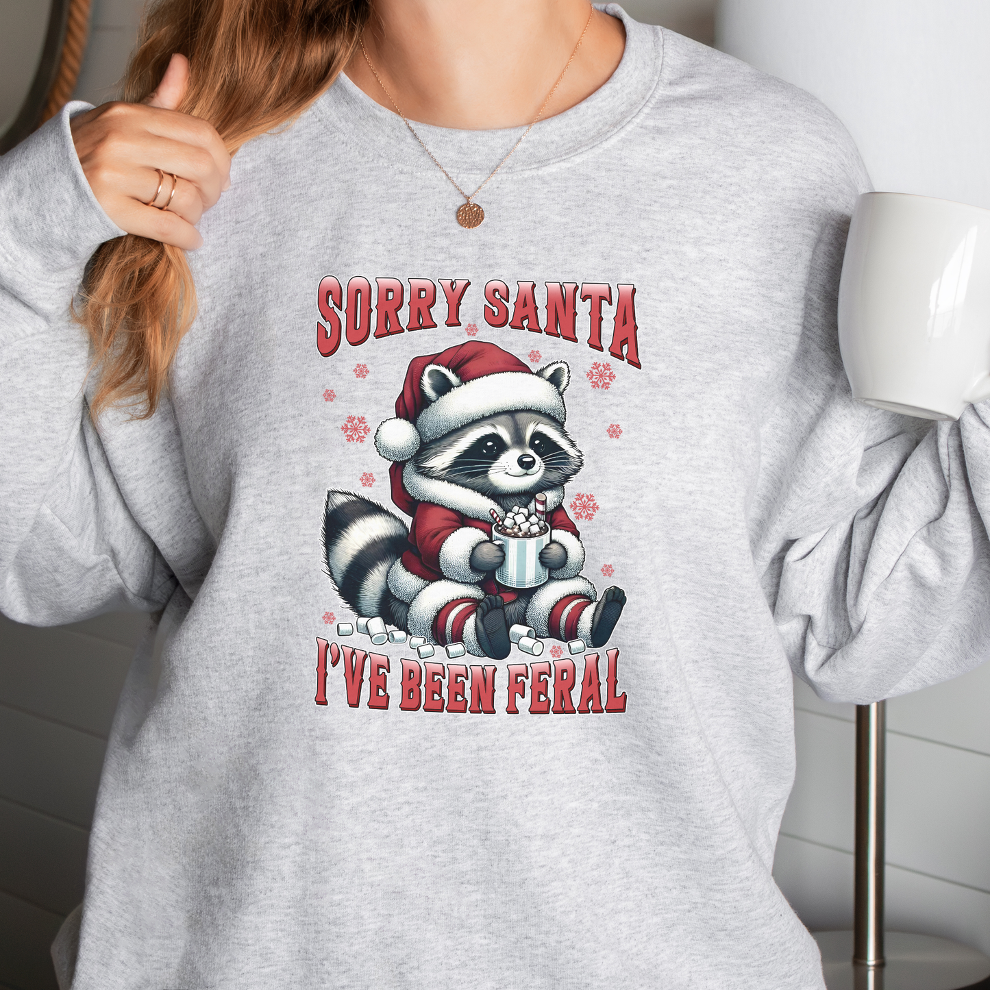 Person wearing a grey sweatshirt with a Christmas-themed  raccoon design in a snowy outdoor setting, sorry Santa I've been feral, 