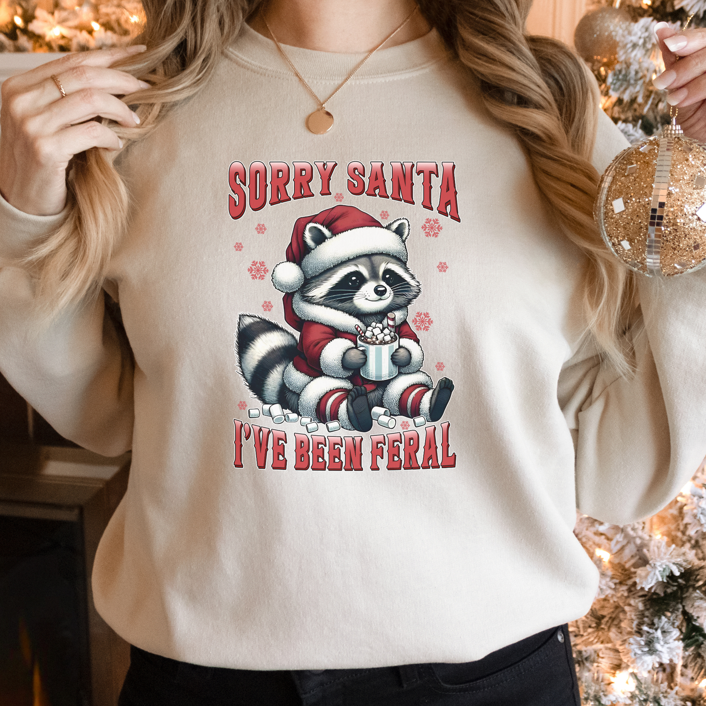 Person wearing a sand sweatshirt with a Christmas-themed  raccoon design in a snowy outdoor setting, sorry Santa I've been feral, 