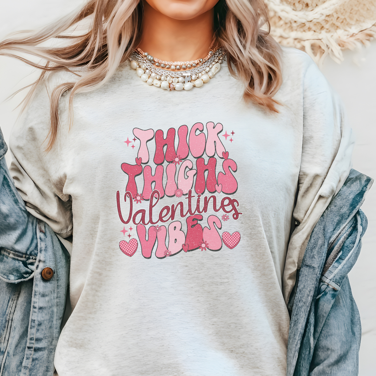 Person wearing a light gray t-shirt with pink text 'THICK THIGHS VALENTINE VIBES' anda  denim jacket.