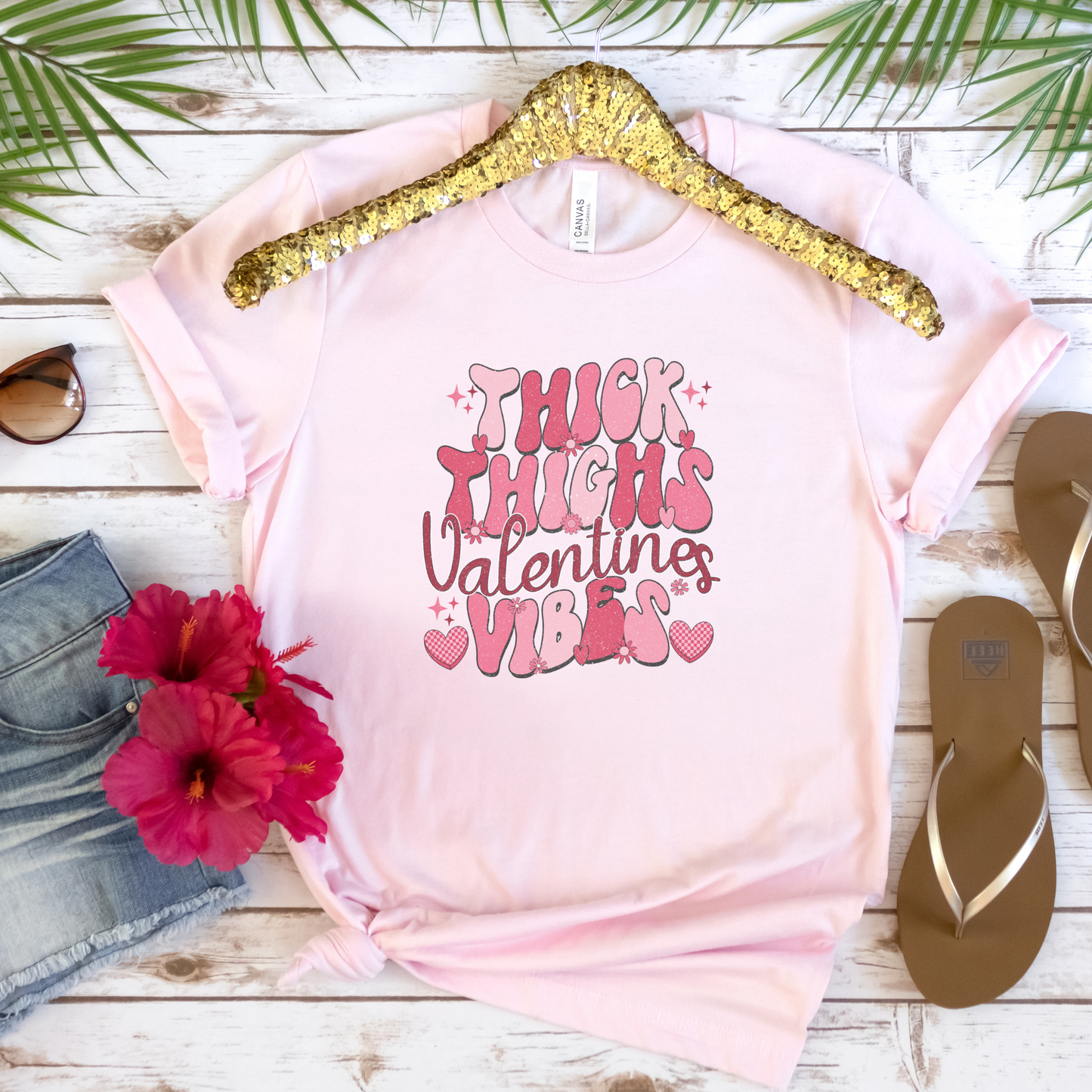 Pink t-shirt with 'Thick Thighs Valentine's Vibes' text on a wooden surface with flowers and sandals.